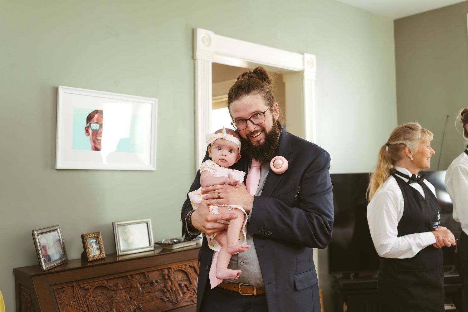 Man with bun hairstyle holds smiling baby in pink outfit at a wedding celebration.