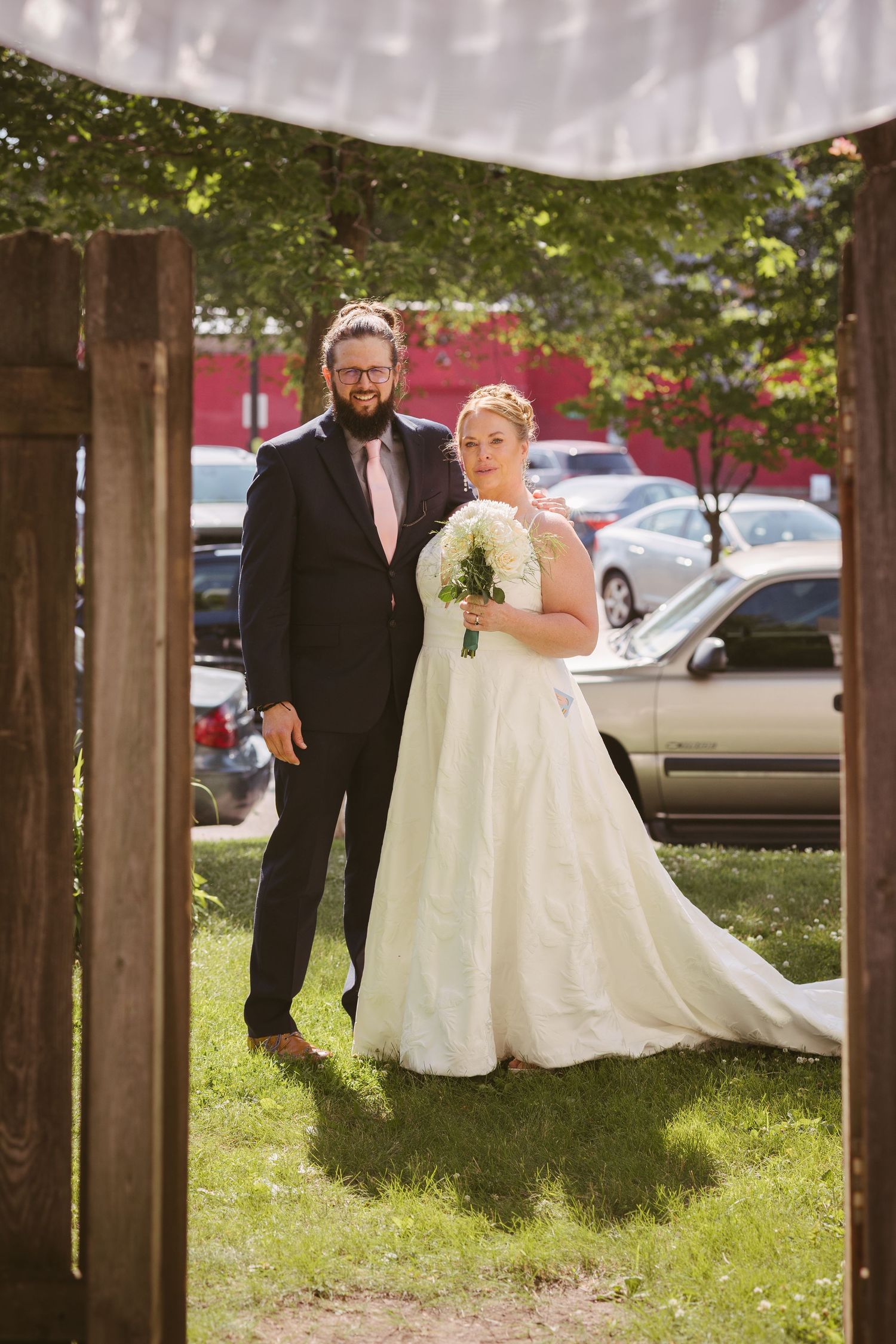 Bride and groom standing together framed by wooden arch, bride holding white bouquet in flowing wedding dress.