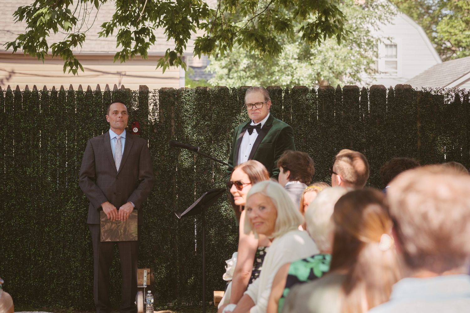 Man in tuxedo officiating outdoor wedding ceremony before seated guests with green hedge backdrop.