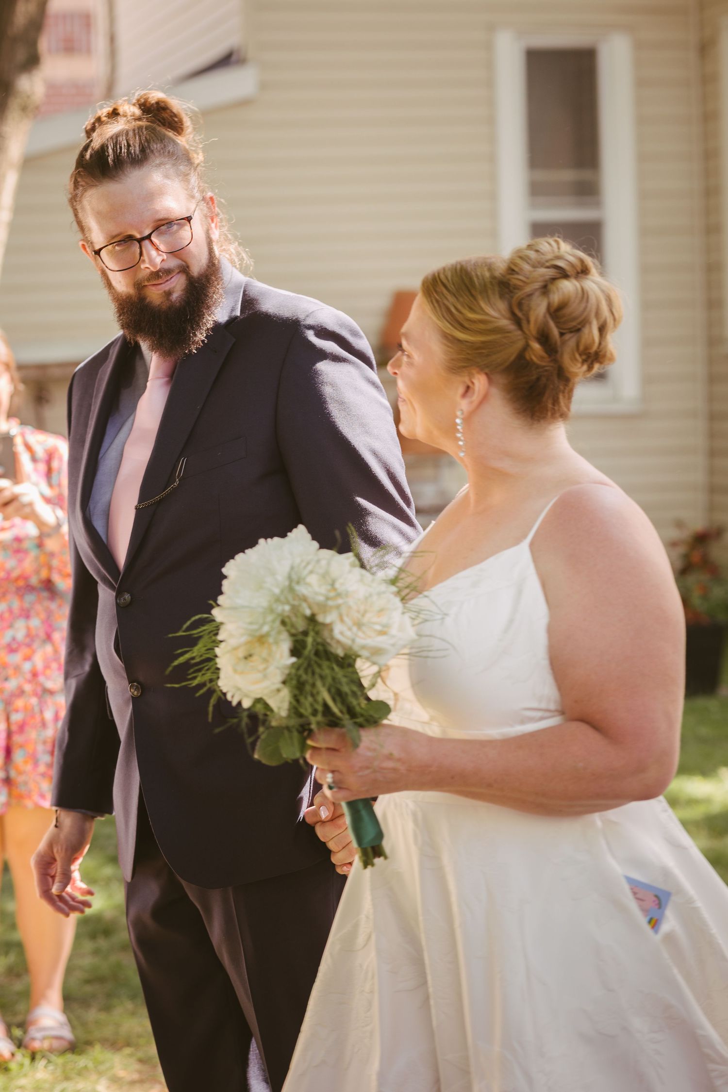 Bearded groom in navy suit walks beside smiling bride holding white bouquet during outdoor wedding ceremony.