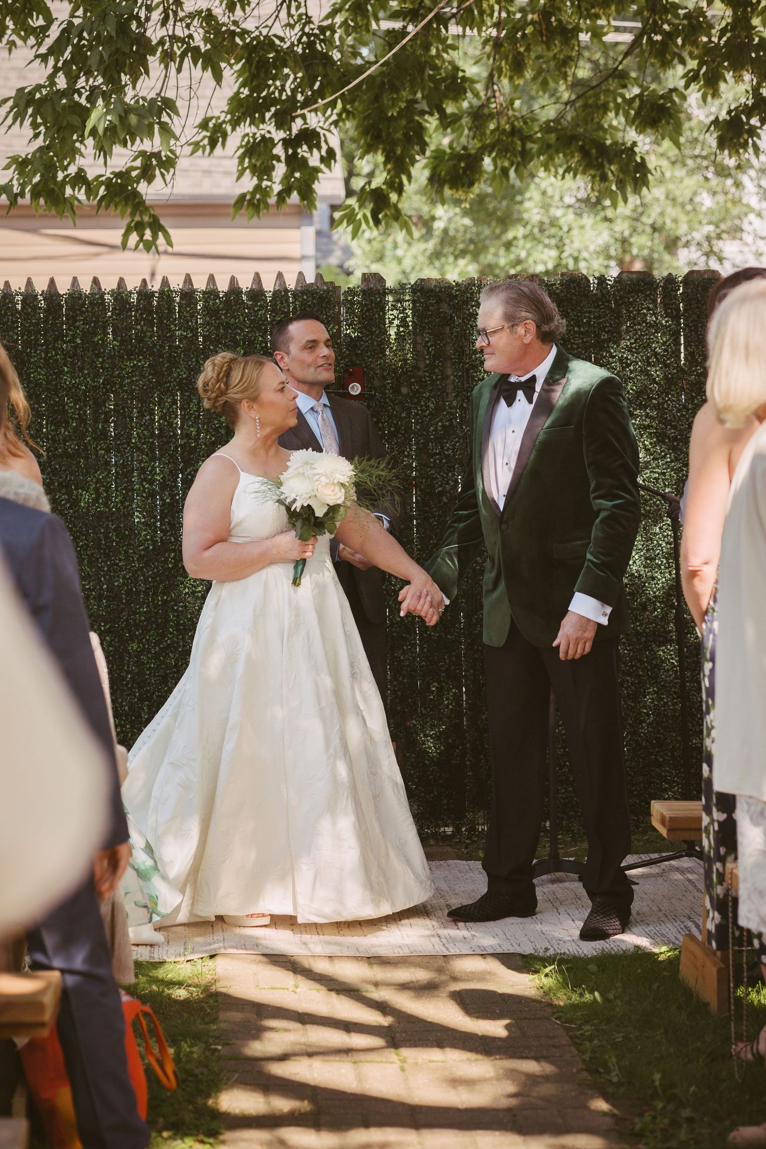 Bride in white gown holding white bouquet walks with man in green velvet tuxedo at outdoor wedding ceremony.