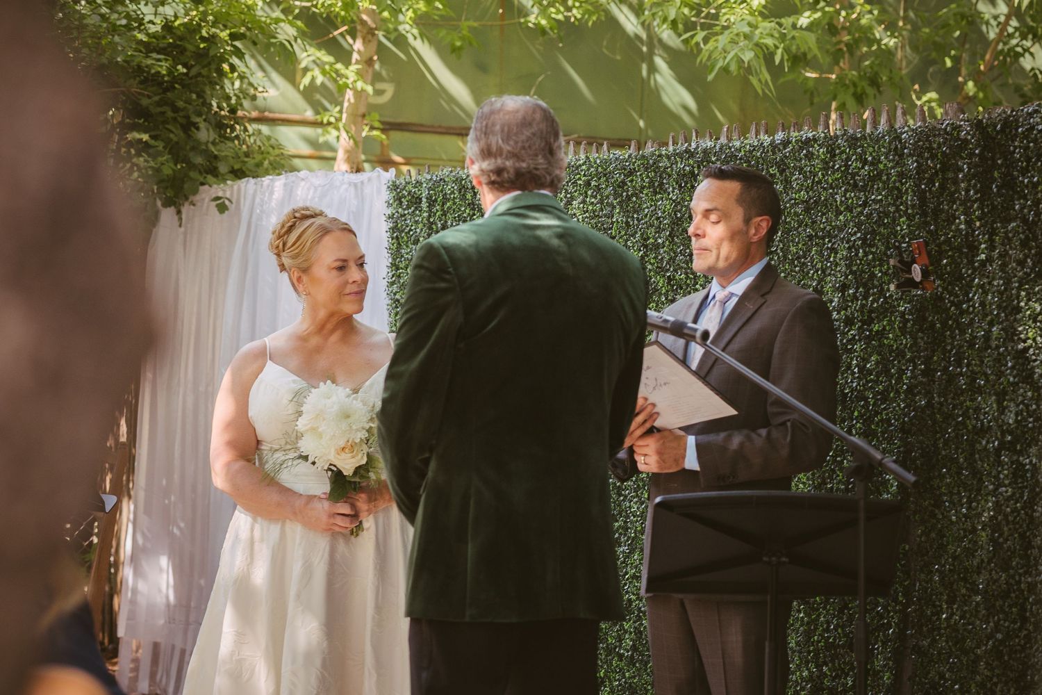 Bride in white gown holds white bouquet during outdoor wedding ceremony with officiant and groom at hedge backdrop.