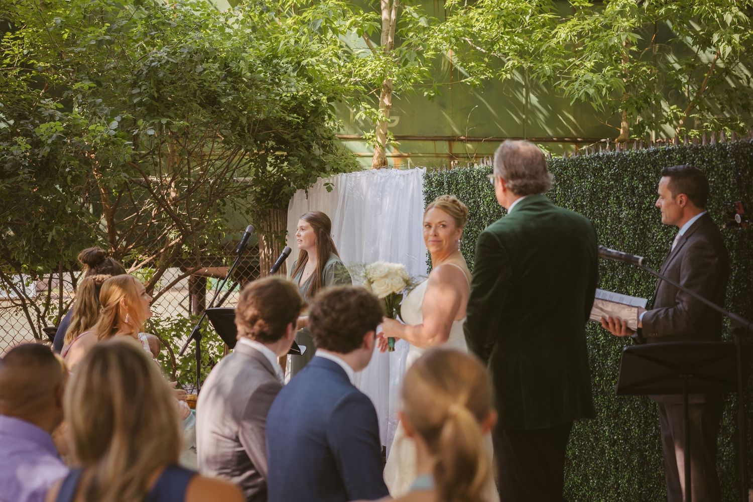 Bride and groom exchange vows at an outdoor wedding ceremony surrounded by lush greenery with seated guests watching.