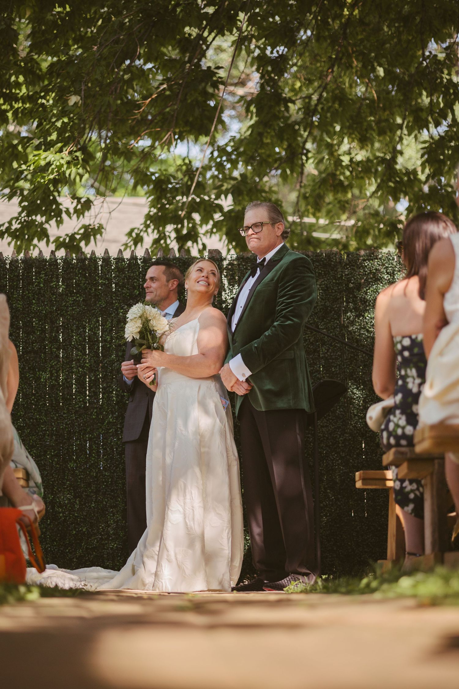 Bride smiling joyfully during outdoor wedding ceremony, standing with officiant beneath lush green trees.