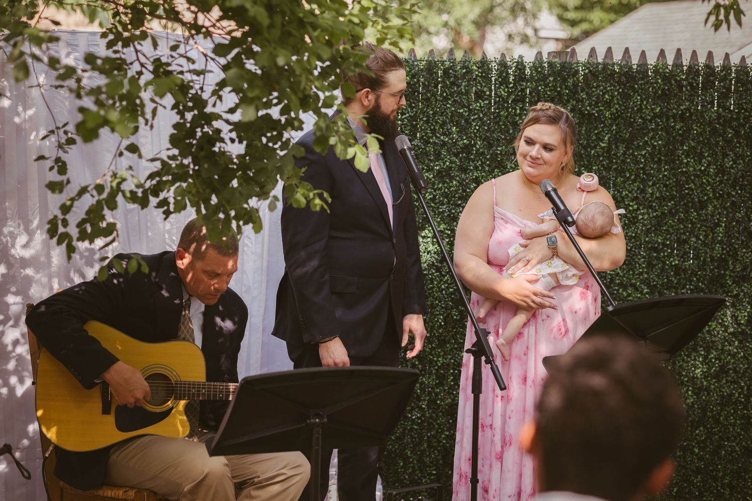 Wedding ceremony with guitarist, groom in suit, and bridesmaid in pink floral dress holding a baby.