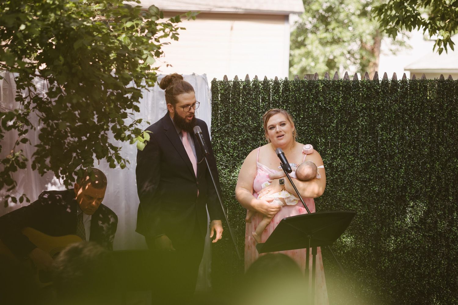 A man in a black suit and woman in a pink dress perform a wedding ceremony reading outdoors.