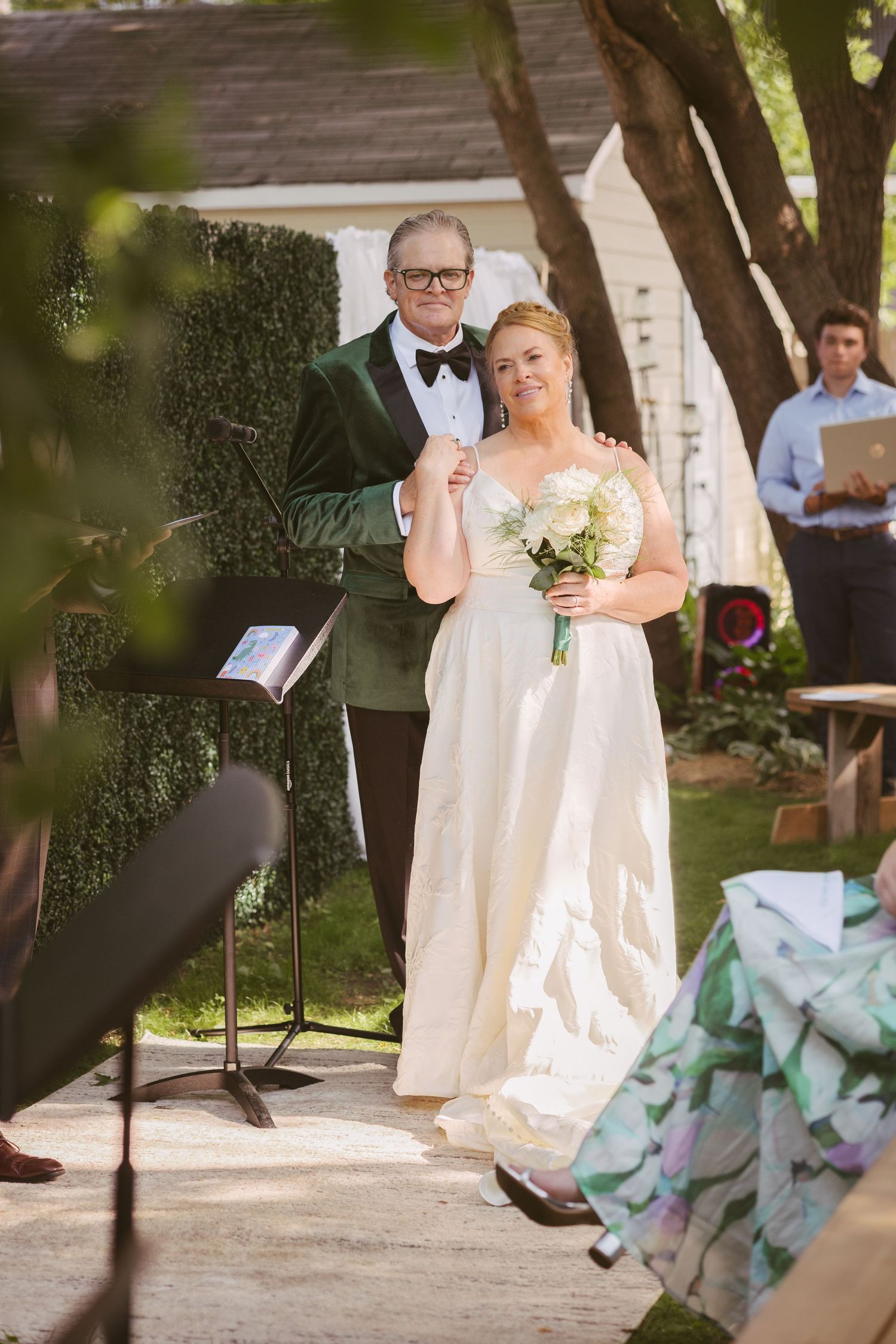 Bride in white gown holds bouquet while walking down outdoor aisle with older man in green tuxedo jacket.