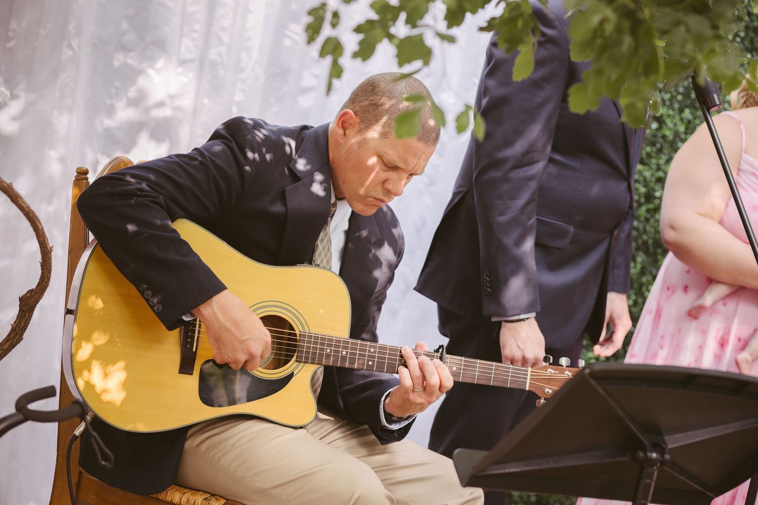 Musician playing acoustic guitar during outdoor wedding ceremony beneath a canopy of trees.