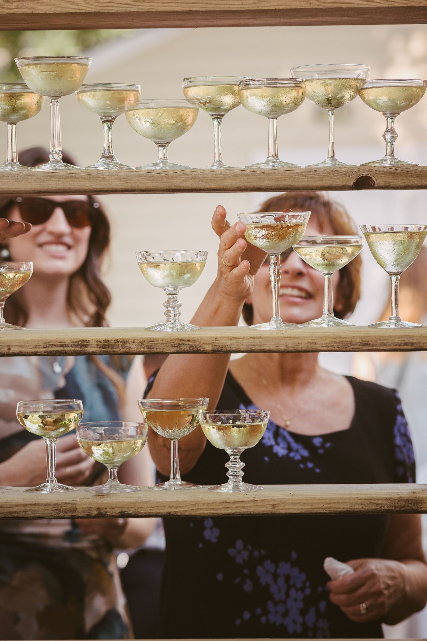 Smiling wedding guests reach for champagne coupe glasses displayed on rustic wooden shelf.
