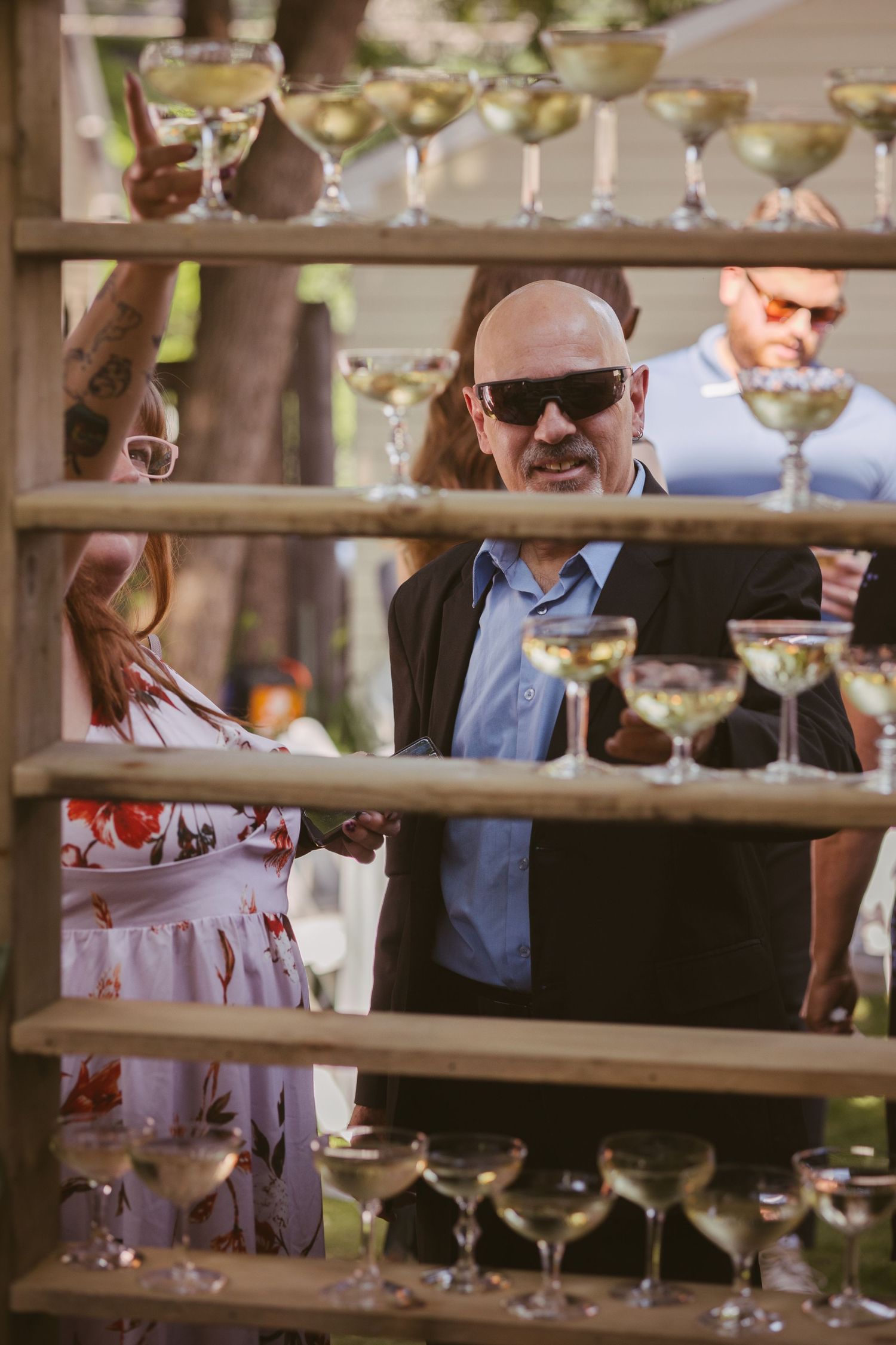 Bald man in sunglasses and woman browse champagne glasses displayed on wooden shelves at event.