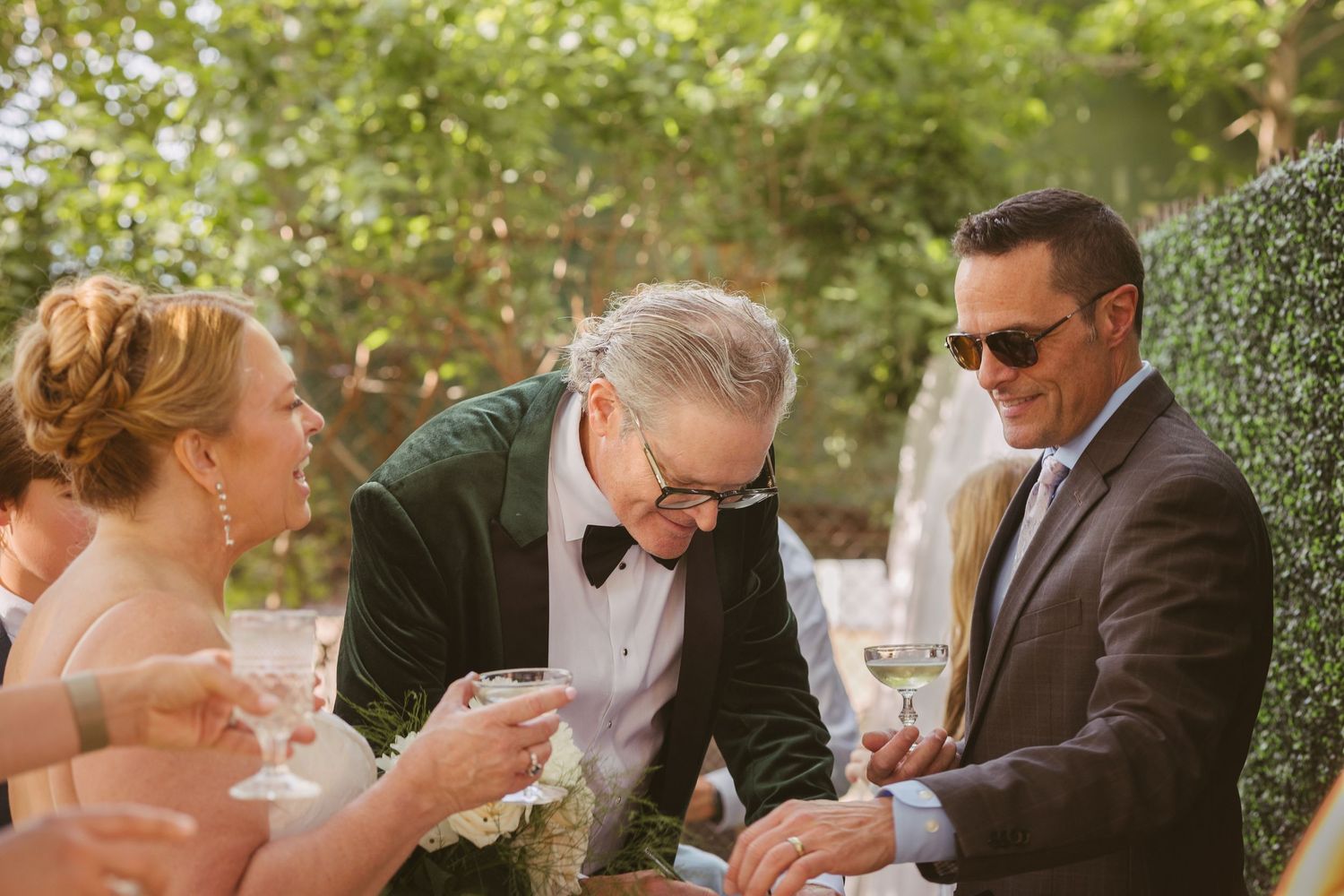 Groom in green velvet tuxedo clinks glasses with guests during outdoor wedding reception in a sunlit garden setting.