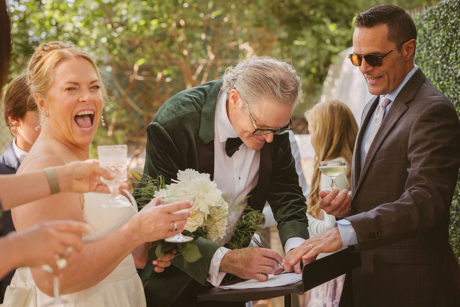 Laughing bride with bouquet and groom in green tuxedo signing wedding documents with a witness.