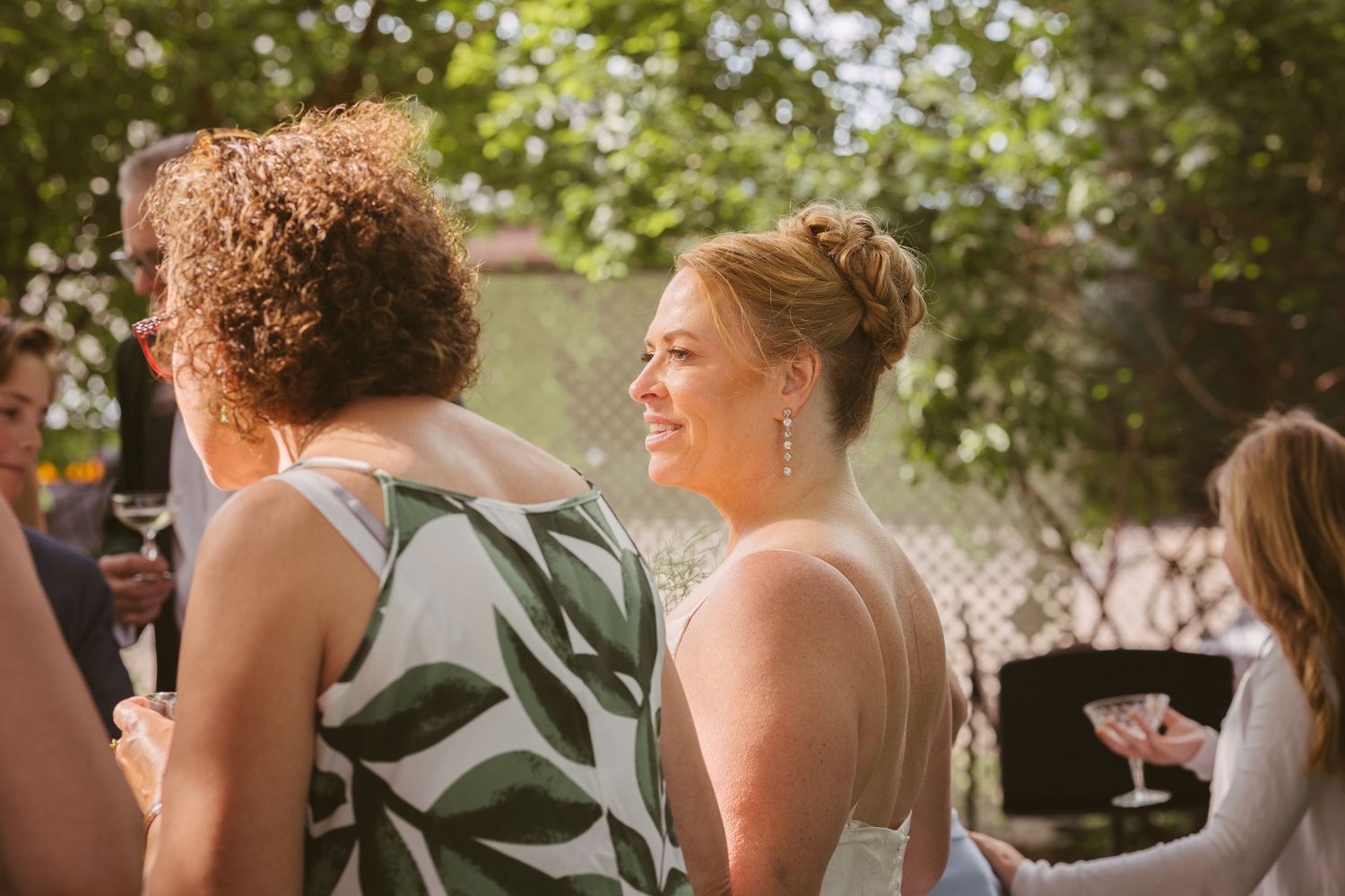 A smiling bride in a white gown chats with guests at an outdoor wedding reception.