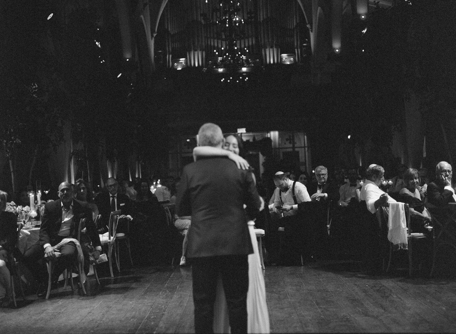 Couple sharing first dance at elegant wedding reception in grand hall with guests seated at tables.