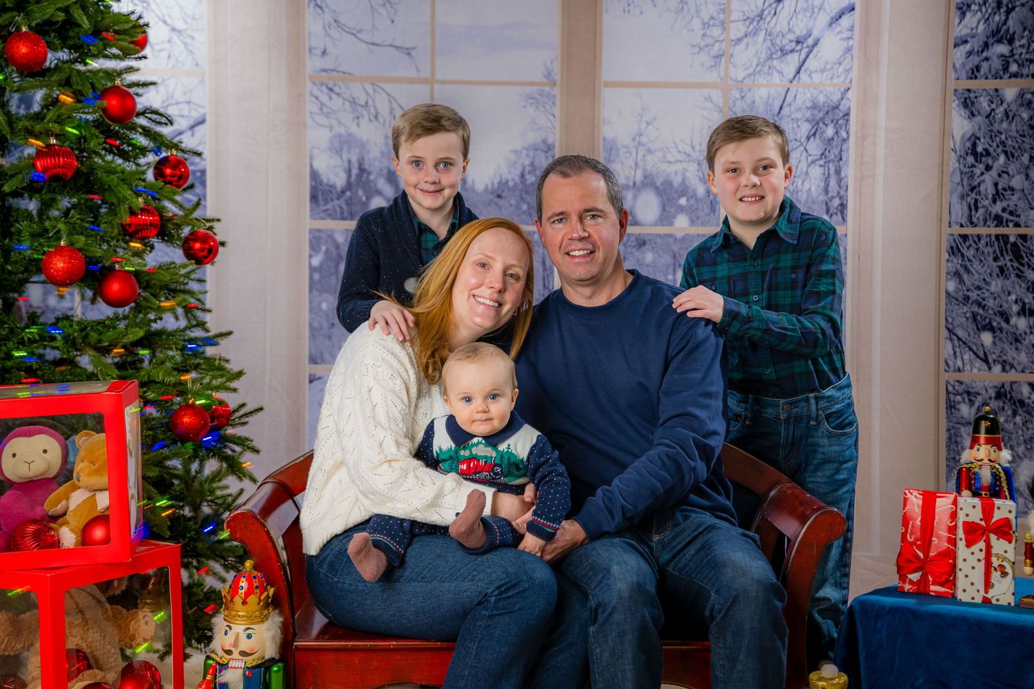 Family of five posing for a Christmas portrait near a decorated tree with gifts and a festive snowy backdrop.