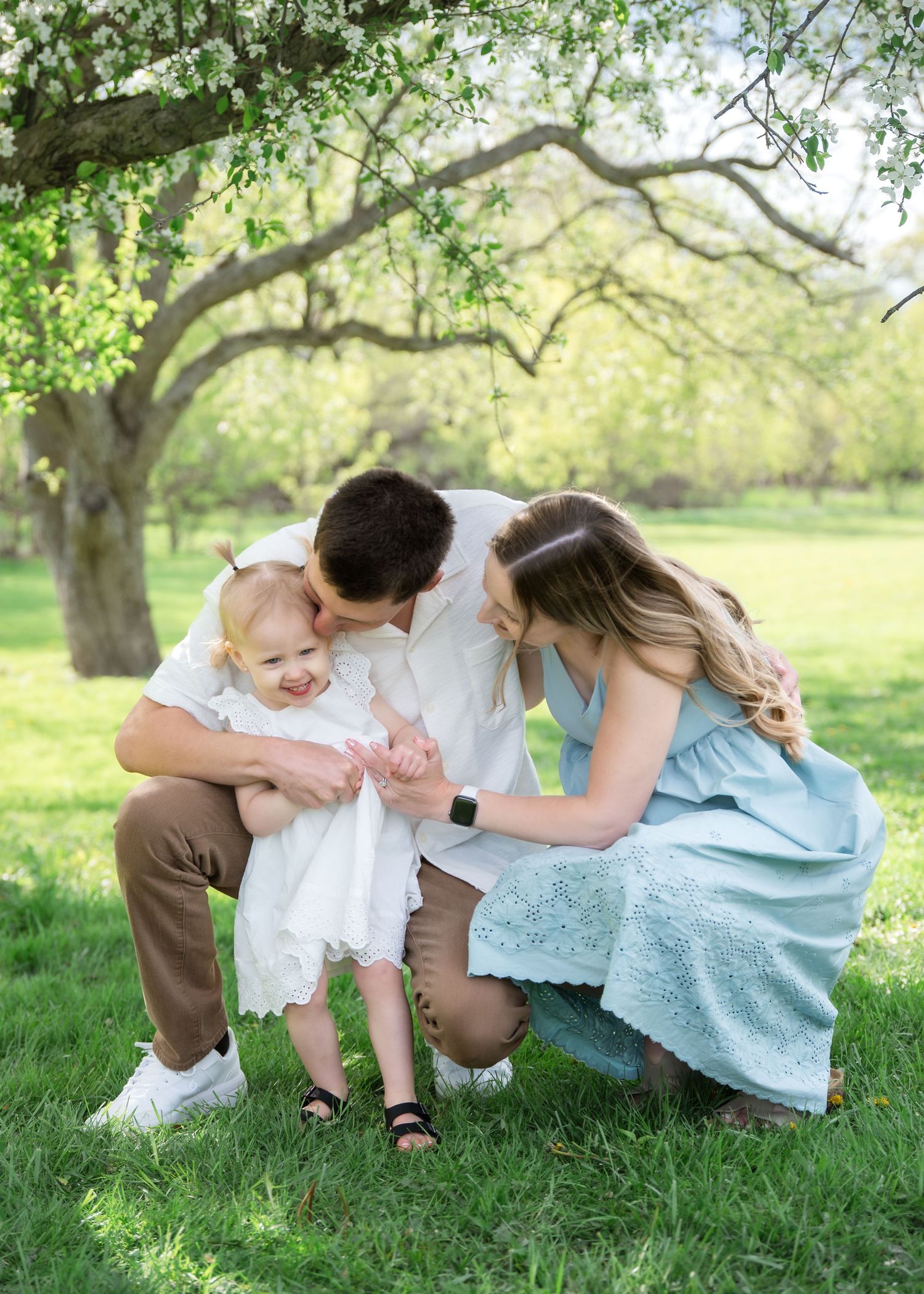 Happy family with toddler playing outdoors in a green park under a tree during a family photo session.