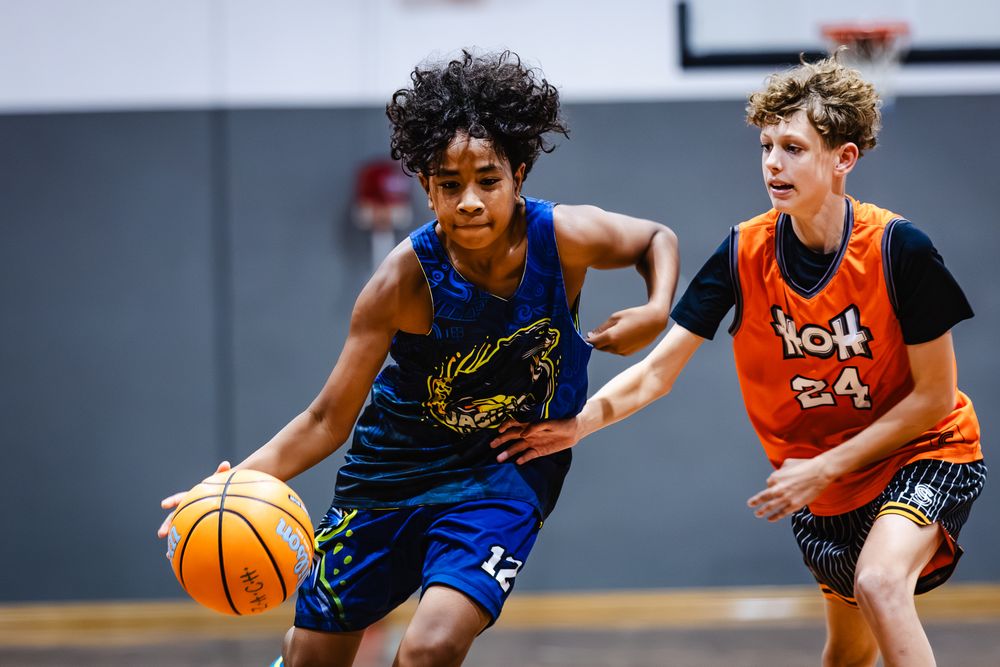Two young basketball players compete on an indoor court, one dribbling in a blue jersey while the other defends in orange.