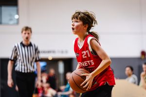 Young girl in red Hornets jersey #27 dribbling basketball during indoor youth game with referee in background.