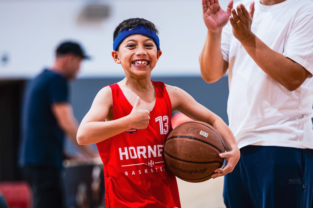 Young boy in red Hornets basketball jersey holding a ball and giving thumbs up at indoor basketball camp.