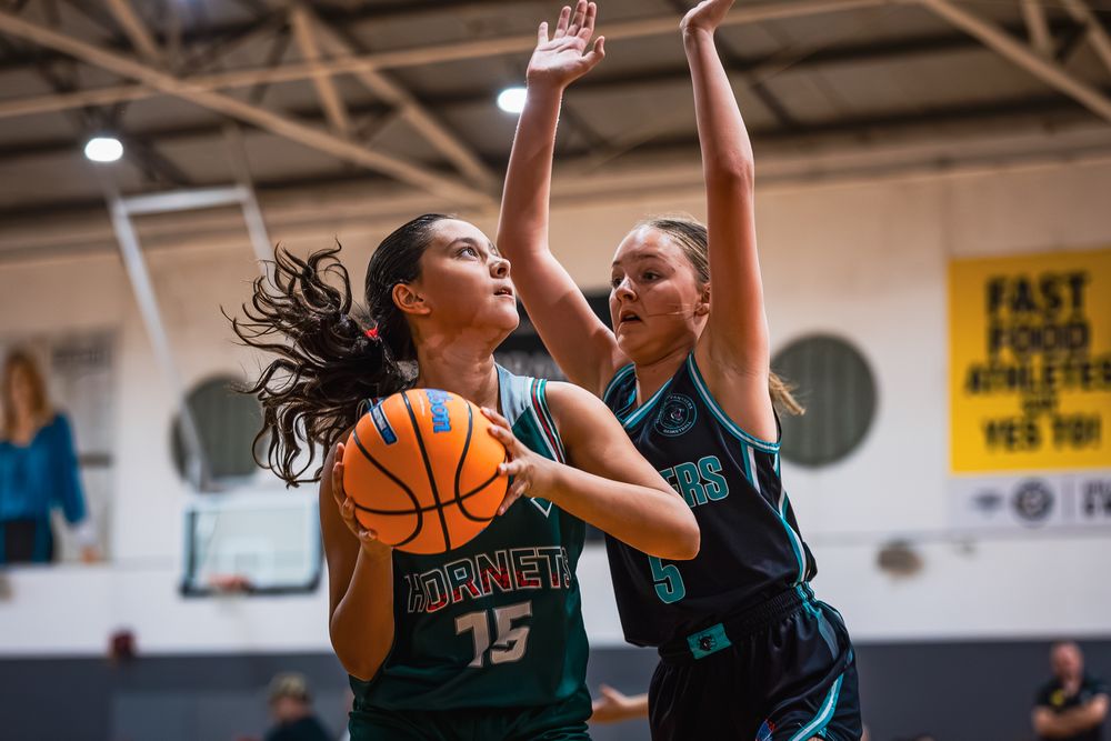 Two female basketball players in teal uniforms compete, with player 15 driving to the basket while defender raises arms to block.