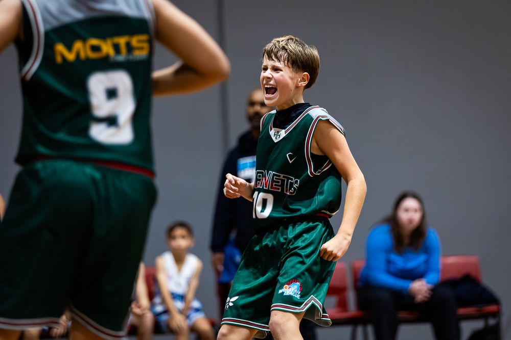 Young basketball player in green jersey celebrates excitedly during youth indoor basketball game.