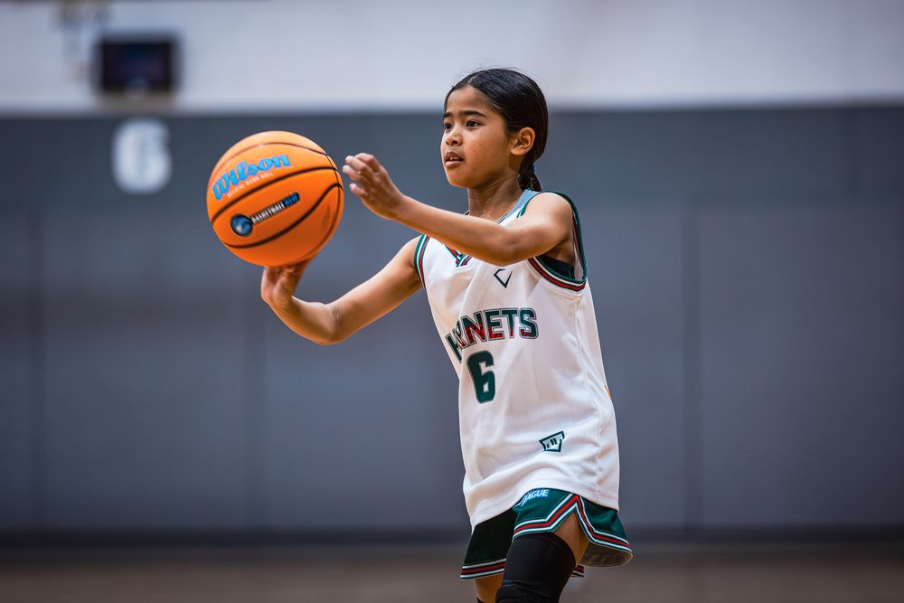 Young girl in Hornets #6 jersey dribbling basketball on indoor court during youth basketball game.