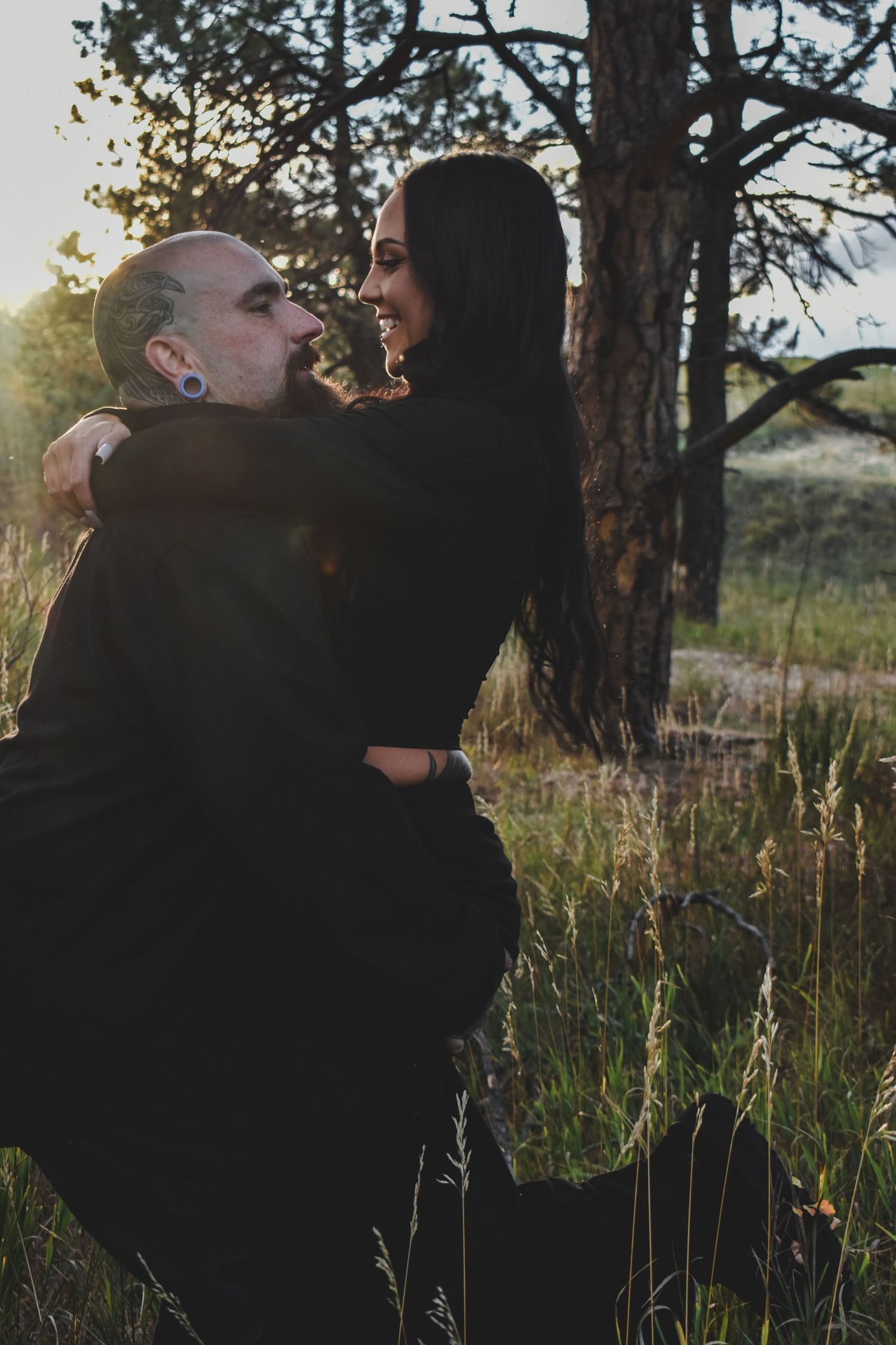 A couple embraces in a sunlit forest meadow, the man kneeling while the woman smiles down at him, both dressed in black.