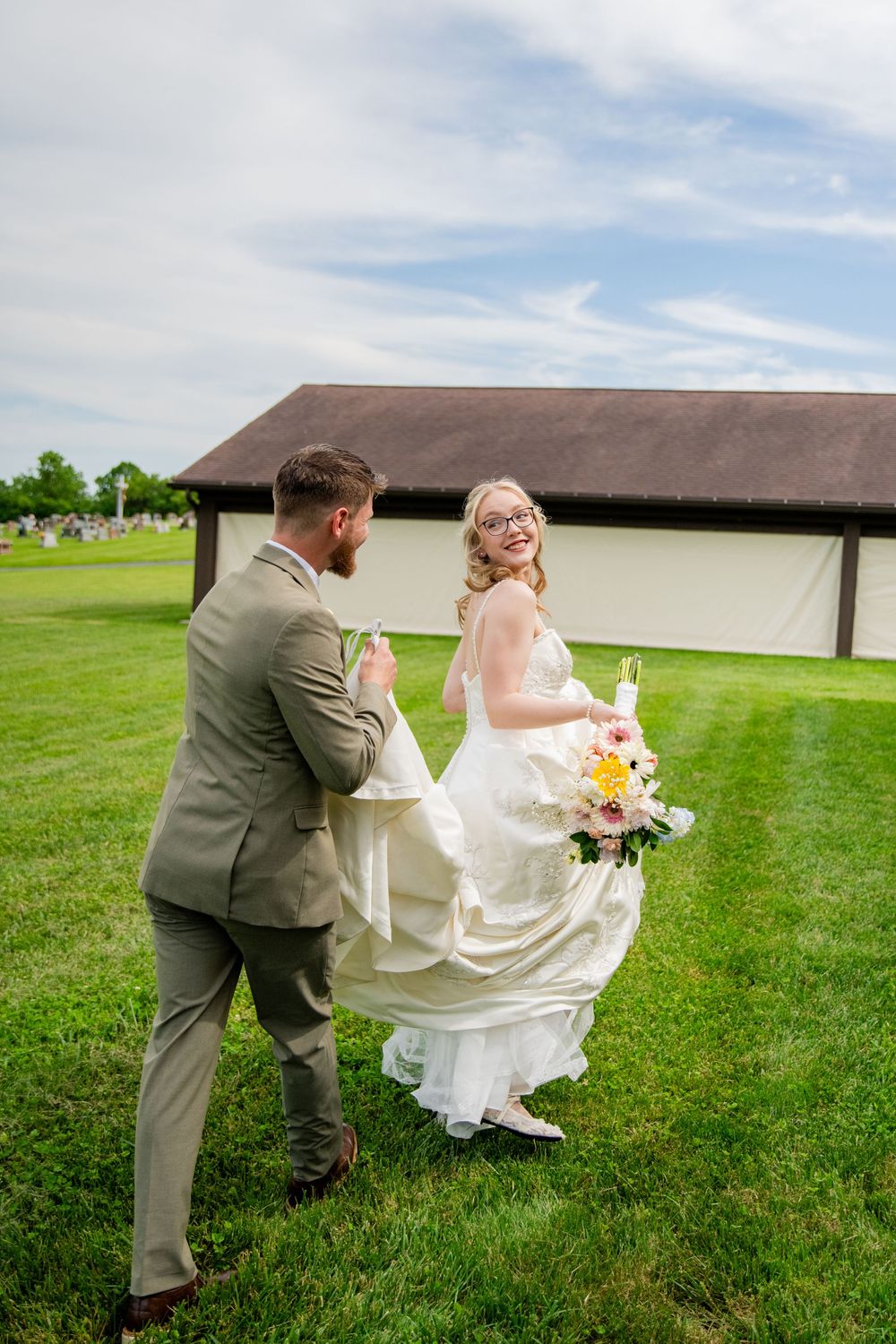 Groom helping smiling bride in white gown walk across green lawn outdoors.