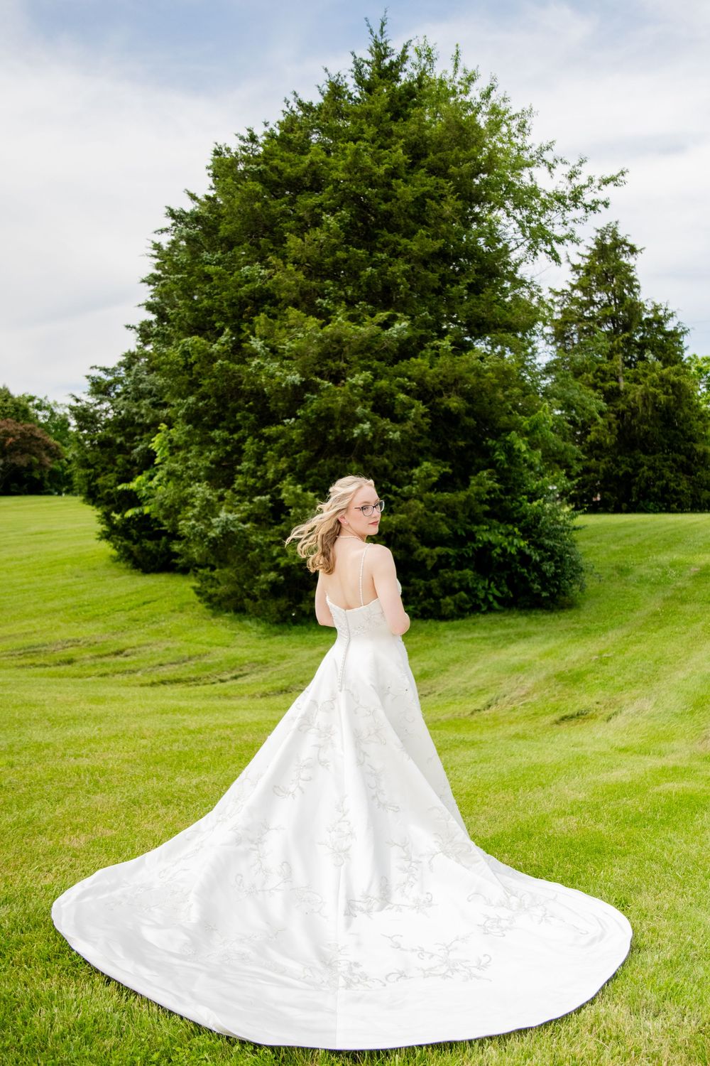 Bride in elegant white gown with long train poses in a lush green park setting.
