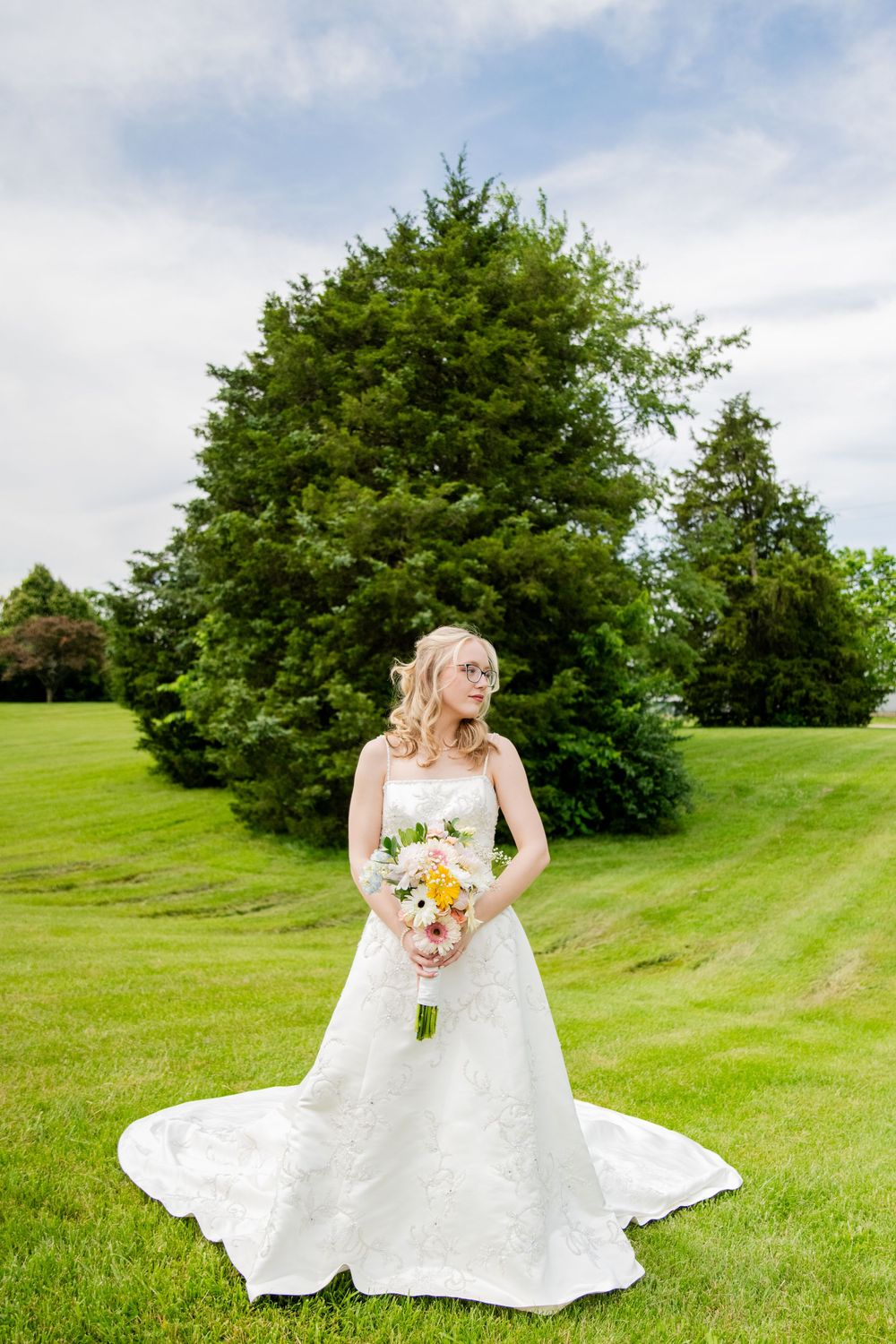 Blonde bride in strapless lace gown holding colorful bouquet standing before evergreen trees.