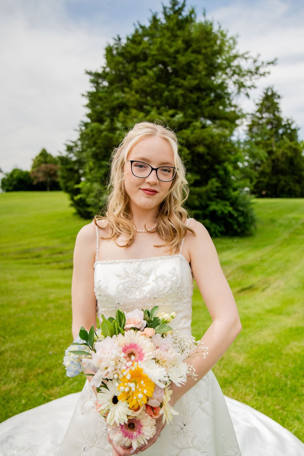 Blonde bride wearing glasses holds colorful bouquet of pink and yellow flowers while posing outdoors on green lawn.