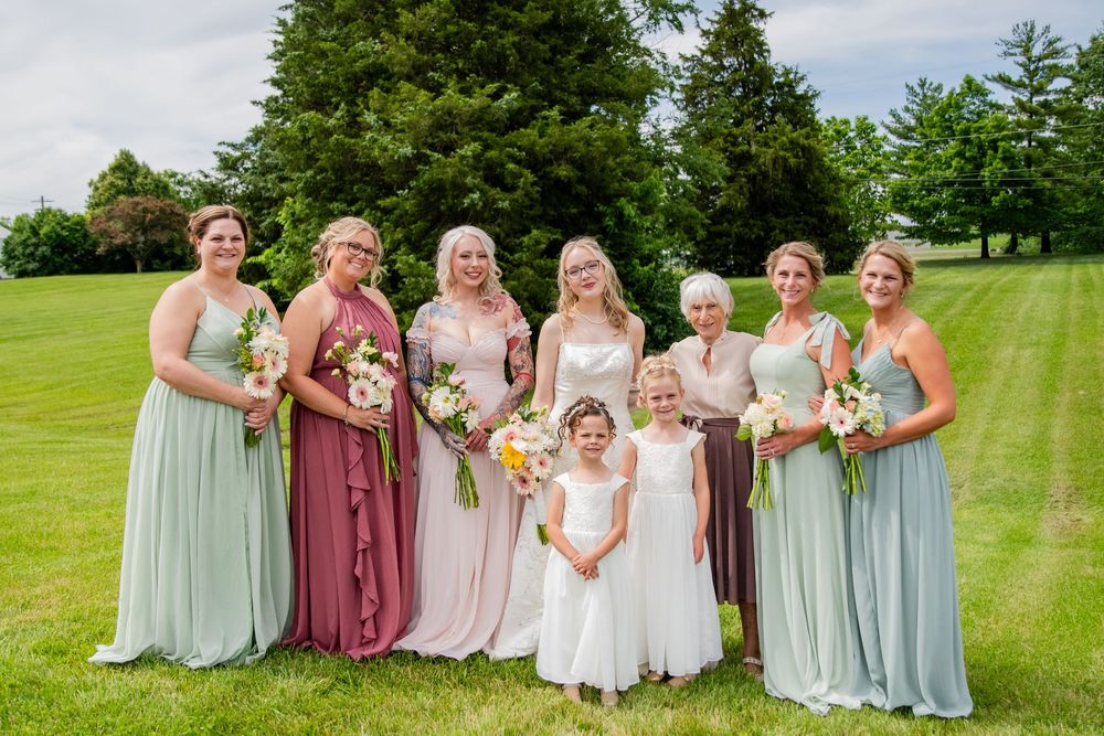 Bridal party in sage and mauve dresses with flower girl poses outdoors on green lawn for group photo.