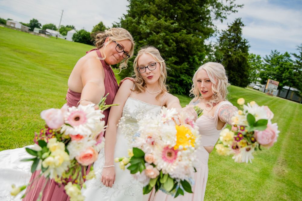 Bride with two bridesmaids in mauve dresses holding vibrant floral bouquets on green lawn.
