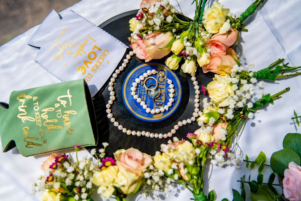 Wedding flat lay featuring pearl necklace, blue brooch, floral bouquets, and gold-lettered koozies on black vinyl record.