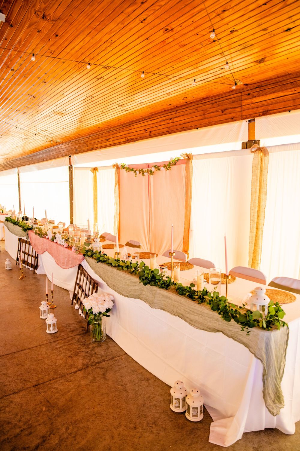 Elegant wedding head table decorated with greenery runner, white flowers, candles, and lanterns in rustic wood venue.