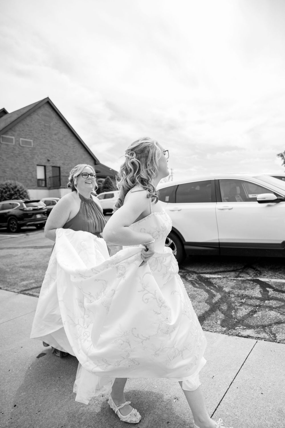 Bride in white gown walks with bridesmaid in black and white photo outside a church before wedding.