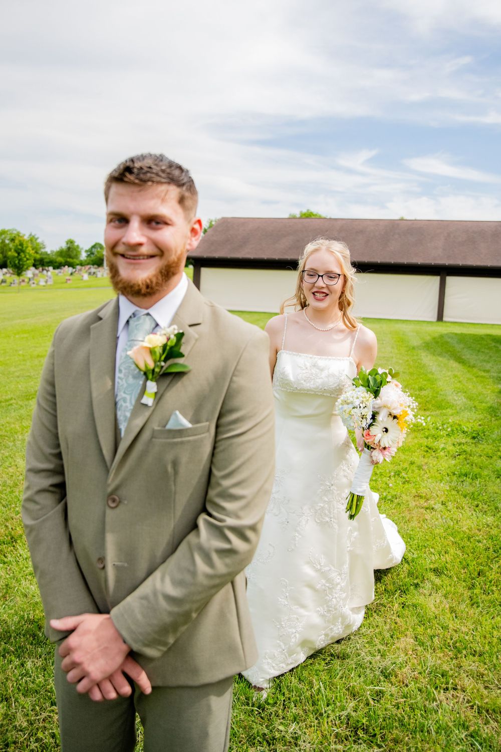Smiling groom in sage suit stands outdoors as bride in white lace gown approaches during first look.