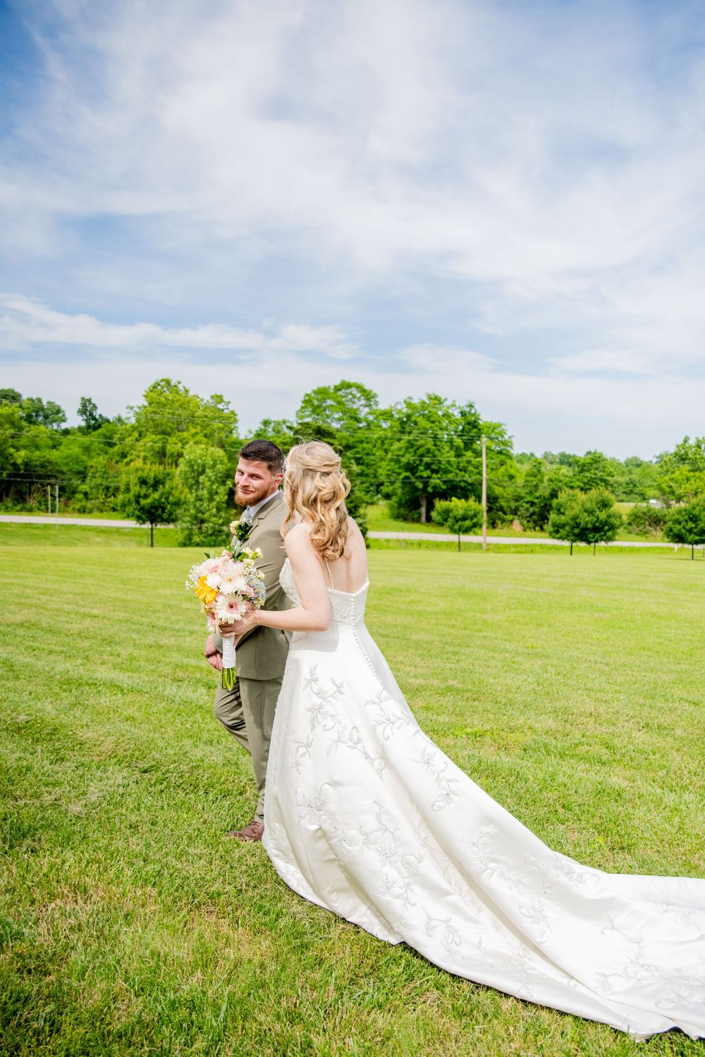 Bride and groom pose back-to-back on green lawn holding floral bouquet with lush trees in background.
