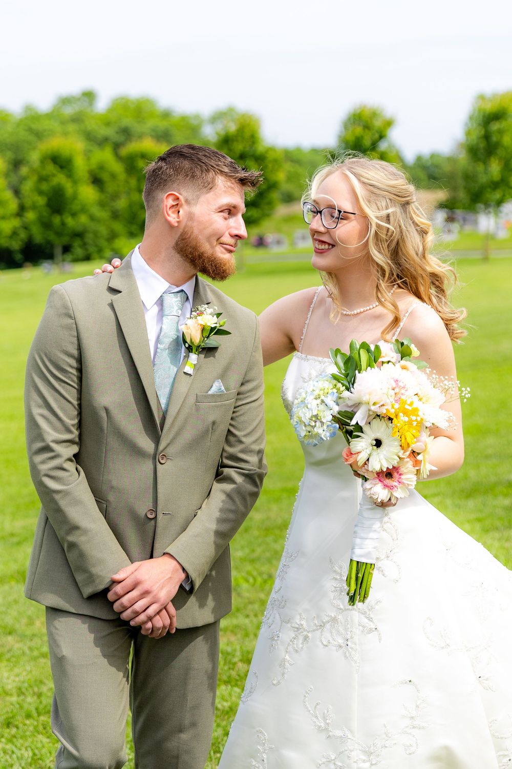 Smiling bride in white gown holding colorful bouquet stands with groom in gray suit on green lawn.