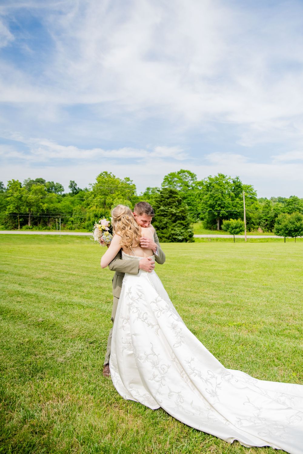 Bride and groom embracing on lush green lawn with bouquet visible against blue sky.