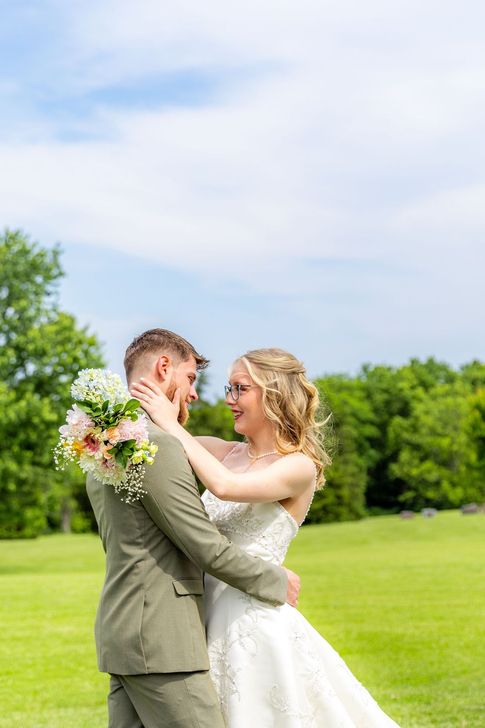 Bride and groom embrace outdoors on a green lawn under a bright blue sky.