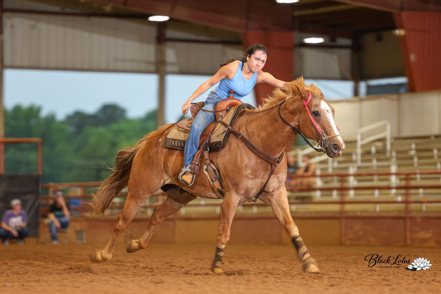 Woman in blue top riding a palomino horse at speed in an indoor rodeo arena competition.