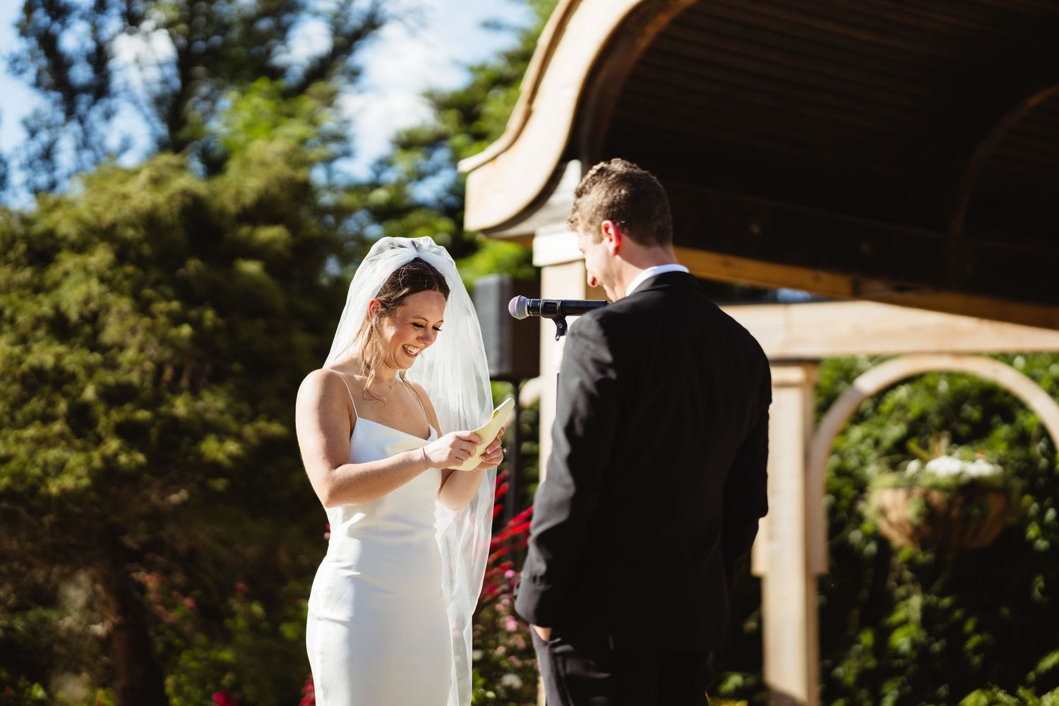 Bride reads vows from paper while groom listens during outdoor garden wedding ceremony near rustic gazebo.