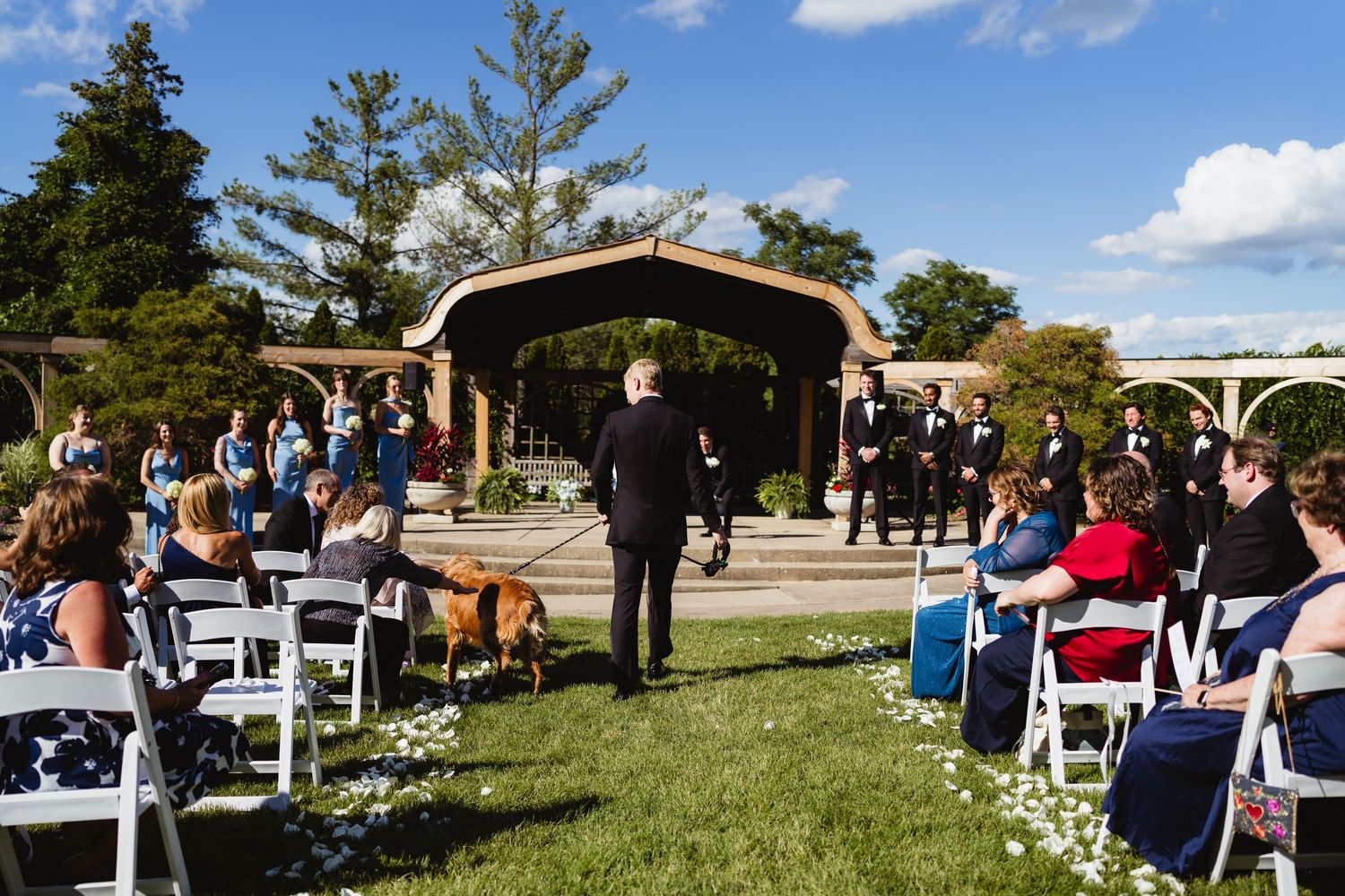 Outdoor wedding ceremony on green lawn with white chairs, a dog in the aisle, and a wooden pergola backdrop.