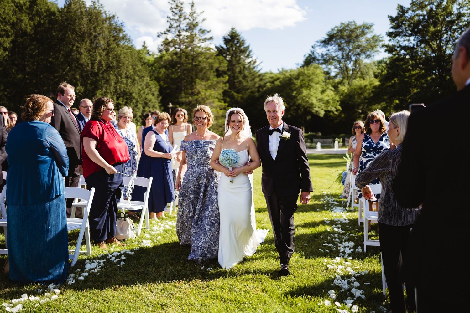 Bride walks down outdoor wedding aisle with parents on sunny day, guests seated on white chairs on green lawn.