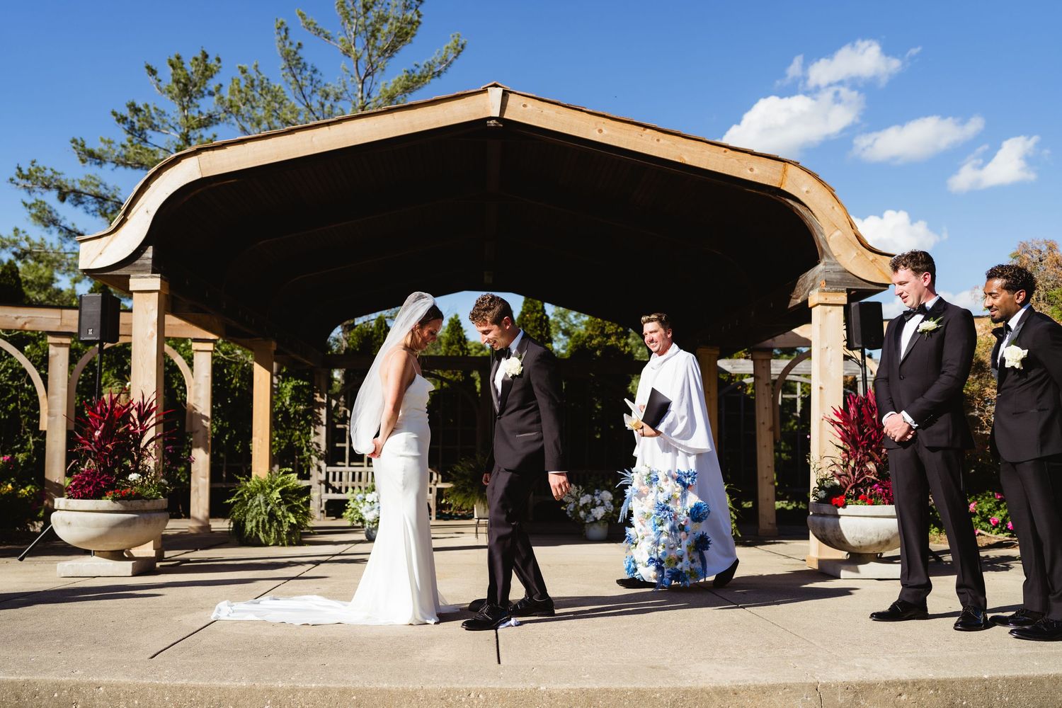 Bride and groom hold hands during outdoor wedding ceremony under elegant pavilion with officiant and groomsman.
