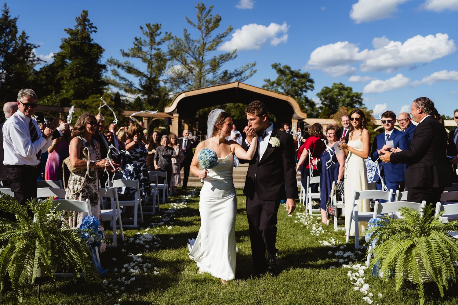 Newlyweds walk down outdoor wedding aisle as guests celebrate under blue sky with rustic wooden arch backdrop.