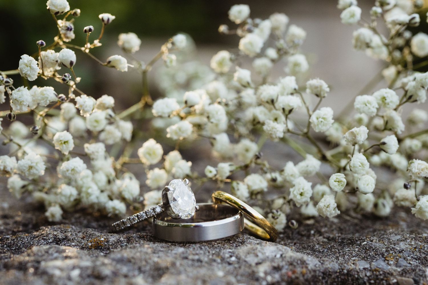 Wedding rings with diamond engagement ring rest on stone surrounded by white baby's breath flowers.