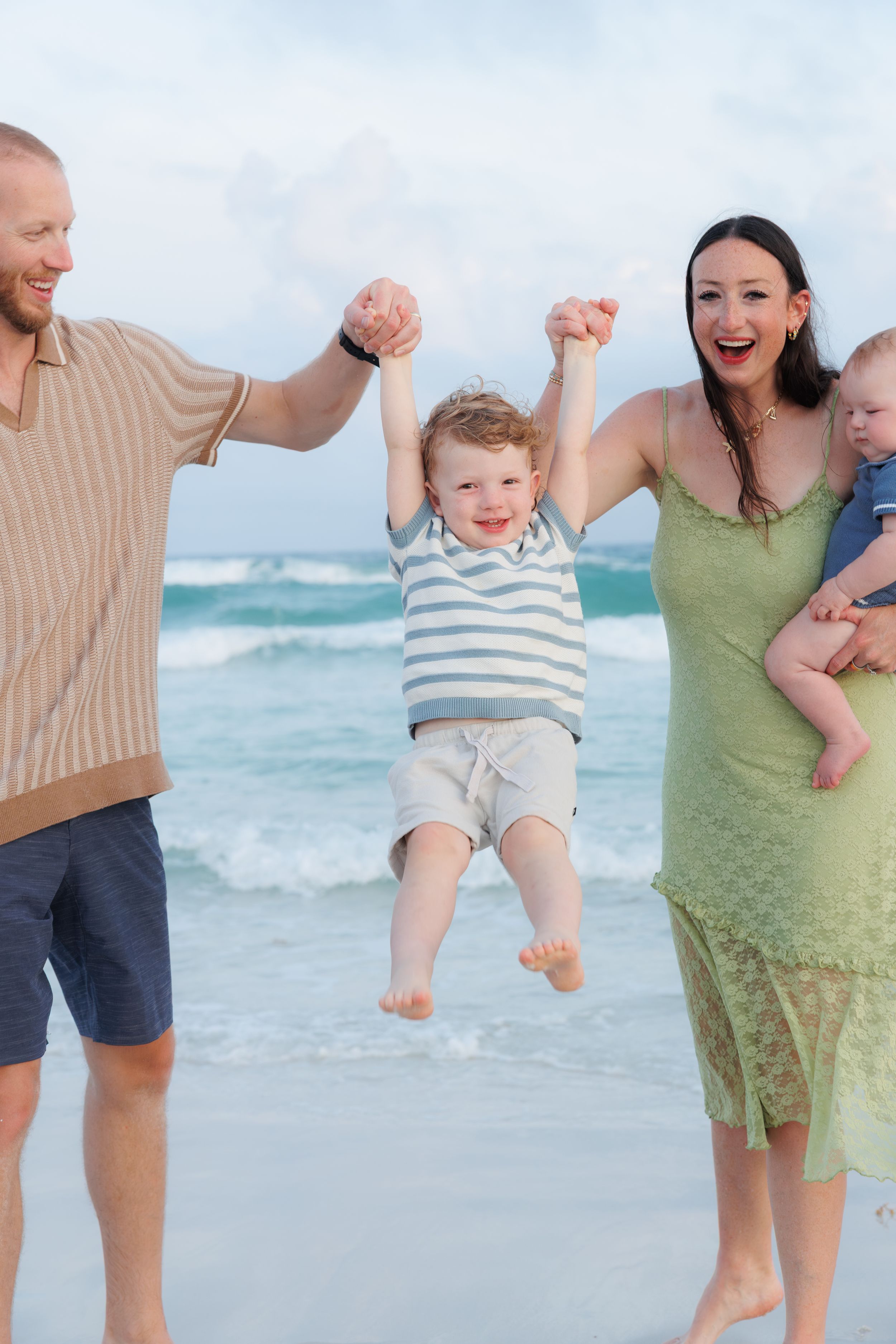 Family at beach, parents swinging toddler by hands while mom holds baby, waves in background.