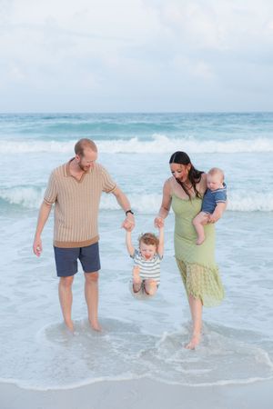 Family of four enjoying a beach walk, parents swinging toddler over waves while mother holds baby.